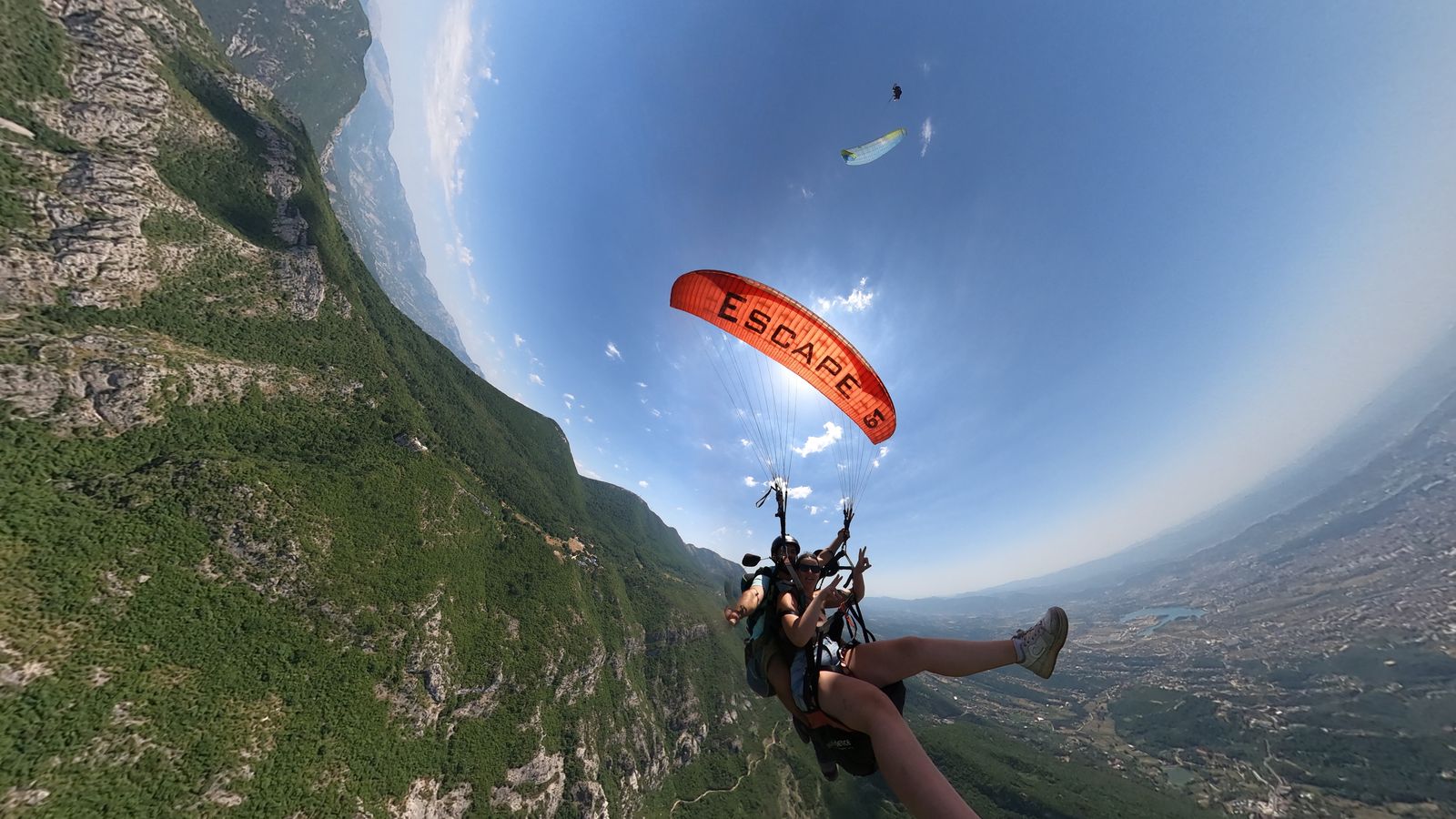Orange paragliding wing backlit by the sun, a second glider higher in the sky, steep wooded slopes below.
