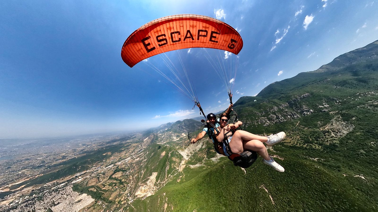Tandem pair under a red paragliding wing, banking over the valley with Tirana's outskirts behind them.