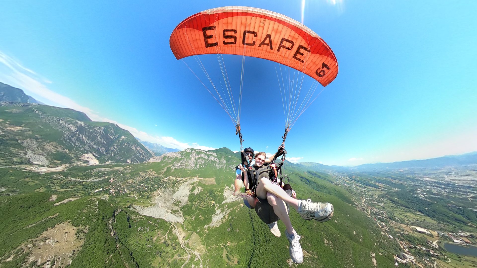 Tandem pair under a red paragliding wing high above the Dajti valley, with Tirana spreading out to the horizon.